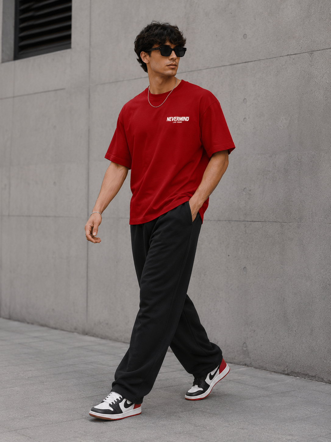 Person wearing a red t-shirt with text, black pants, and black and white sneakers against a gray wall.